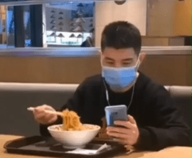A man at a restaurant holding his phone in one hand and chopsticks in the other, focused on his phone as he picks up noodles, wearing a face mask.