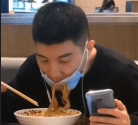 A man in a restaurant attempting to eat noodles while distracted by his mobile phone, still wearing his face mask as the noodles hit the mask.