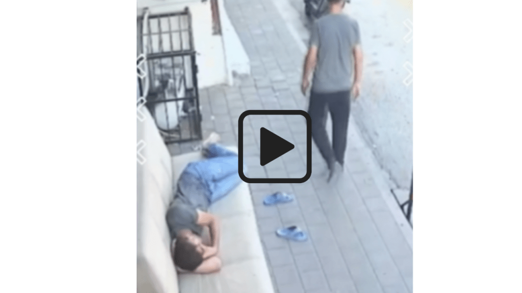 Video of a man sleeping on a sofa on the pavement with a pedestrian nearby.