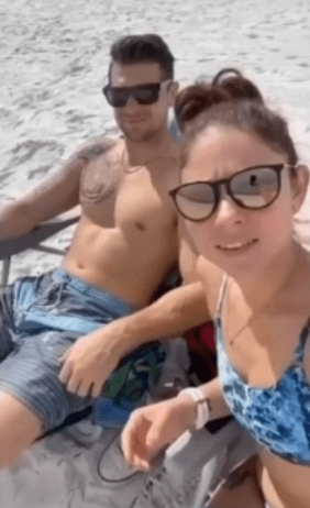 A couple posing for a beach selfie, with the woman playfully wiping sand off her partner's swim trunks.