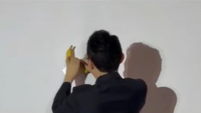 Justin Sun reaches to remove a duct‑taped banana from a white gallery wall