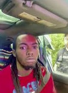 Young Black man seated in driver’s seat of SUV looking toward camera before window is broken.
