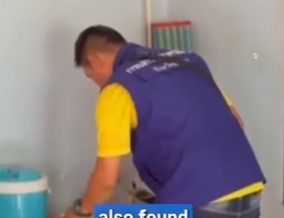 Two uniformed Thai police officers examine a spread of clear plastic drug bags, meth pills, and a homemade handgun on a wooden table