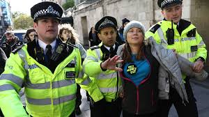 Climate activist Greta Thunberg sits on the pavement during her arrest in London, flanked by two police officers in uniform, with a third in the background, amid a protest setting with metal barriers and a building entrance.