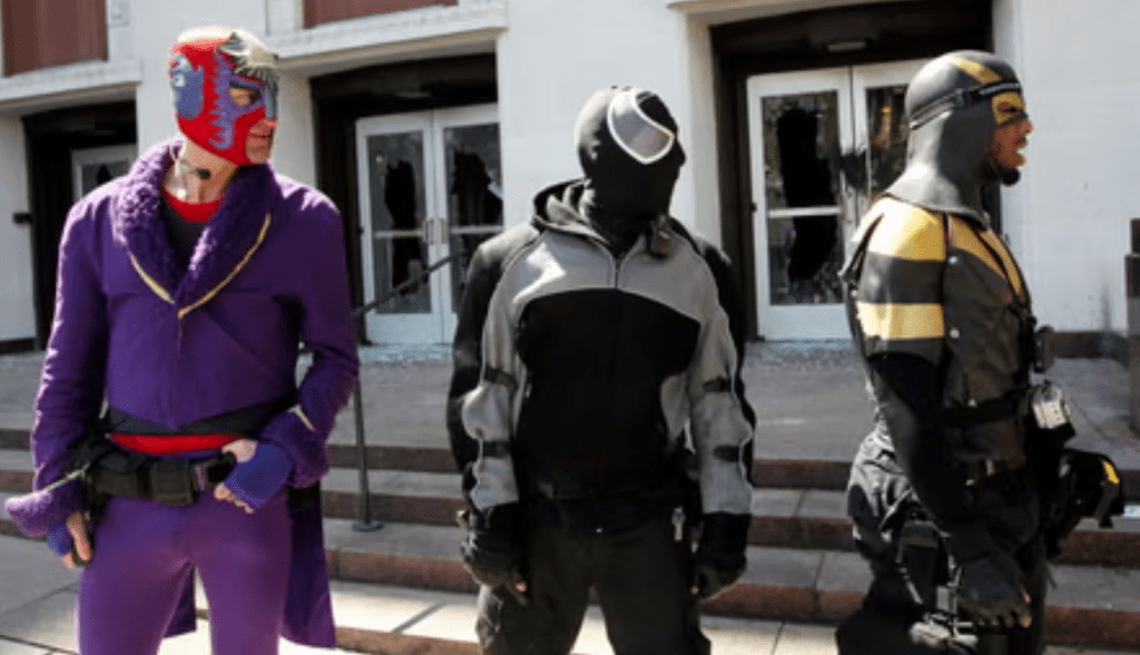 A costumed man wearing a black and gold superhero suit with body armor stands in a city street at night in Seattle.
