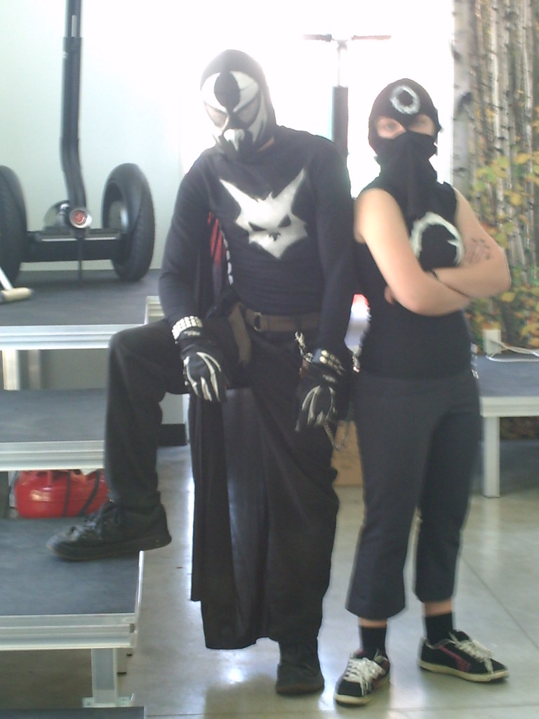 A masked man in a black outfit with a hare symbol stands outdoors, wearing gloves and protective gear.