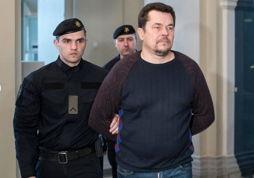 Evaldas Rimasauskas stands handcuffed in a courtroom, flanked by law enforcement officers during a court appearance.