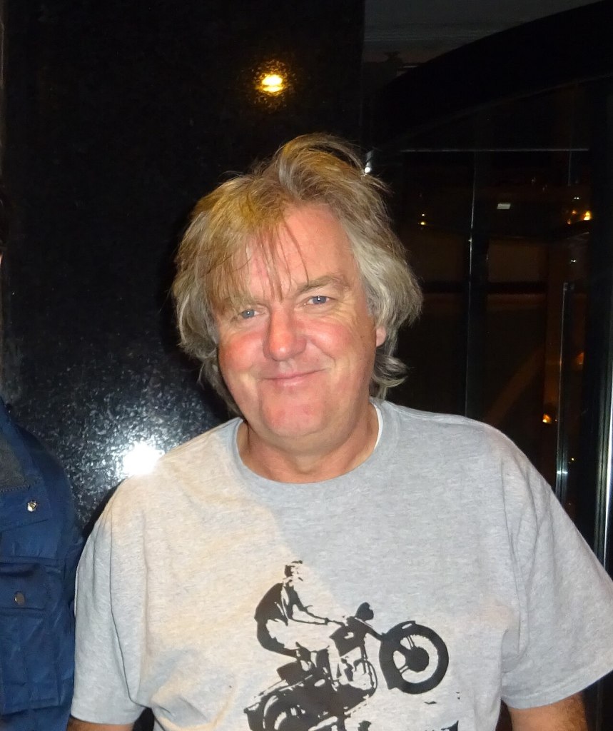 James May smiling at a public event, wearing a jacket and standing indoors.