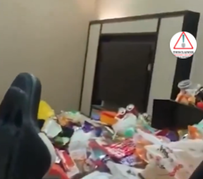 A gaming chair facing a desk in a cluttered room filled with piles of trash, bottles, and food packaging