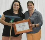 A woman smiling while holding a diploma and a cleaning brush during a staged photo moment.