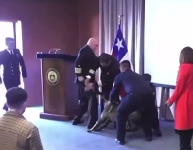 Screenshot of a trained detection dog biting or lunging at a uniformed naval officer during a formal ceremony
