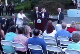 A cyclist rides directly across a formal outdoor ceremony as the seated audience turns to watch in confusion.